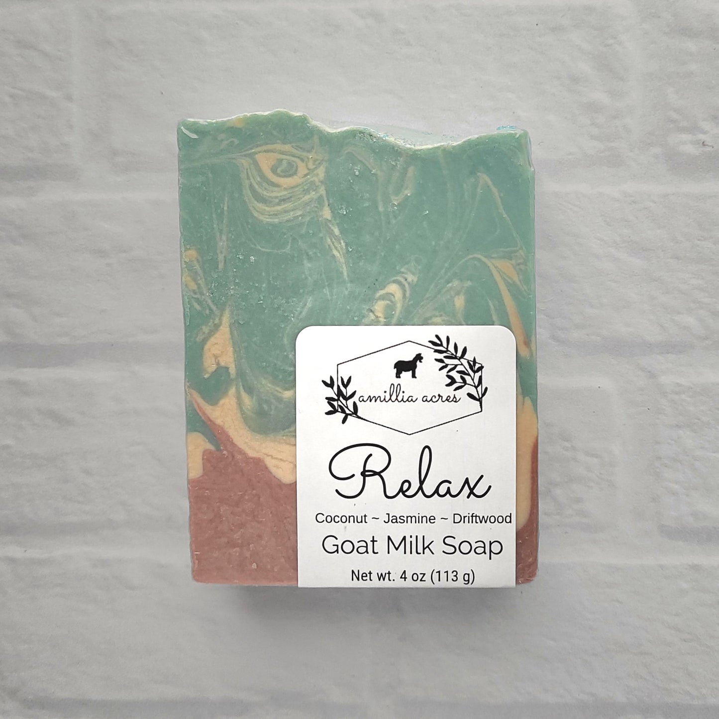 Displayed on a light textured surface, a bar of goat milk soap in green, cream, and brown tones is enriched with natural oils. The label reads: "Amillia Acres ~ Relax | Goat Milk Soap - Coconut - Jasmine - Driftwood - Net wt. 4 oz (113 g)," beautifully showcasing handmade craftsmanship.