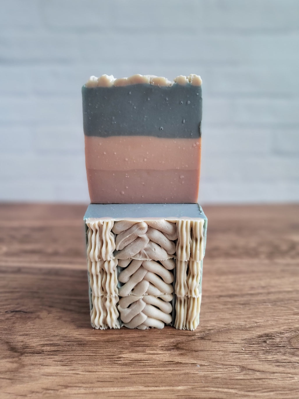 The Sweater Weather goat milk soap from Amillia Acres features intricate gray and pink swirls and braids, displayed atop another soap made from natural oils. The warm wooden surface contrasts against a blurred white brick wall backdrop.