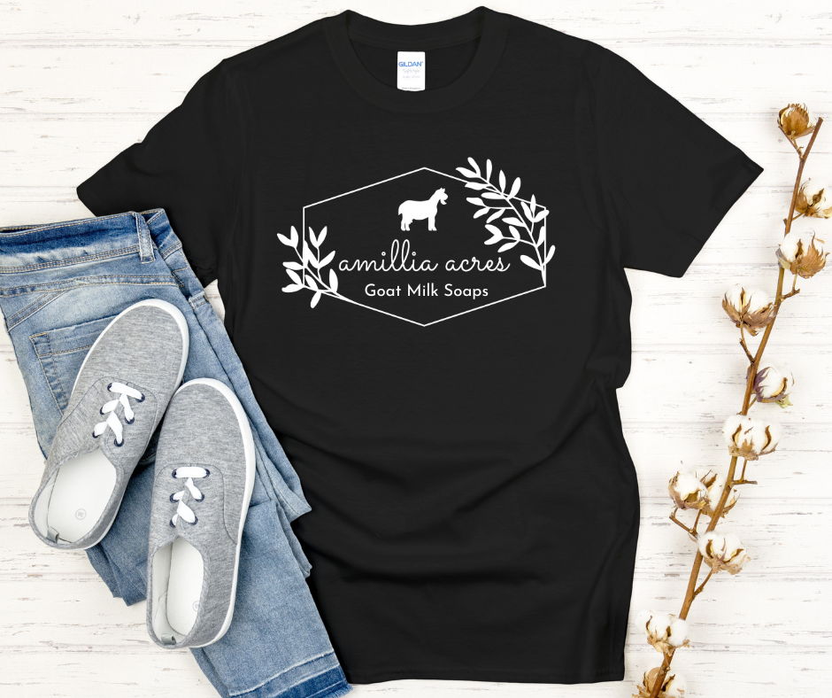 A short sleeve t-shirt by Amillia Acres ~ Goat Milk Soaps and Lotions displays a white goat graphic amidst leaves with "Goat Milk Soaps" text. Made from Gildan Softstyle 100% preshrunk cotton in black, it pairs effortlessly with blue jeans and gray sneakers, complemented by cotton stems nearby.