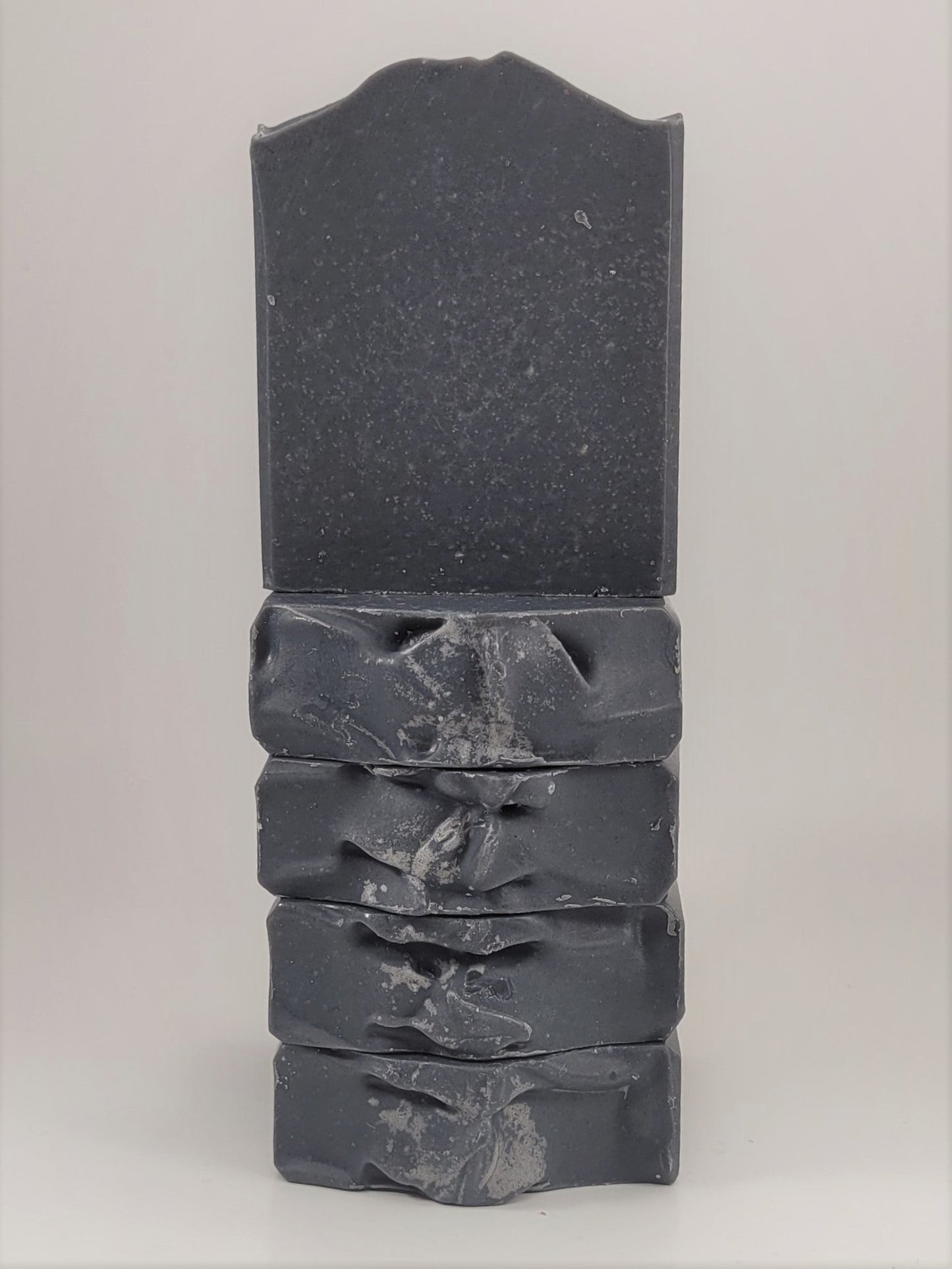 Five dark gray Balance goat milk soap bars by Amillia Acres are stacked on a plain light background. The top bar stands upright, highlighting its rough edges, while the others lie flat below it.