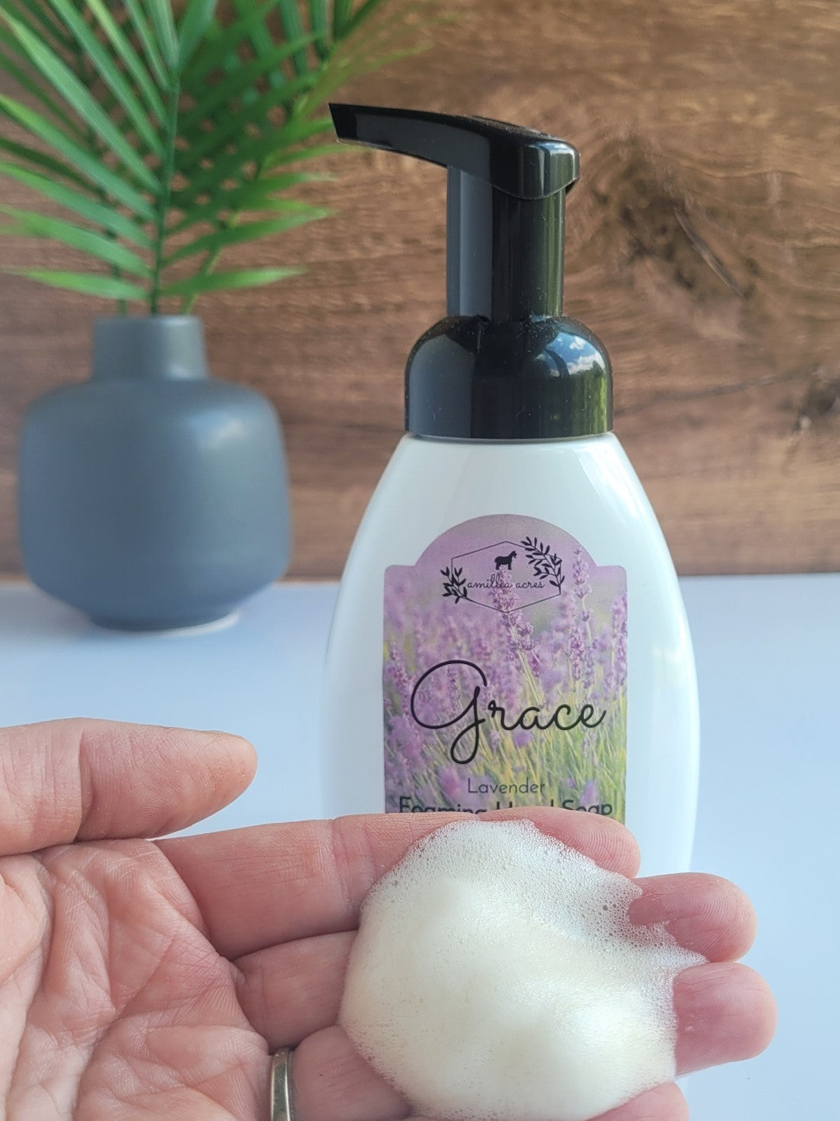 A hand holds foamy soap in front of a "Grace Foaming Hand Soap" bottle by Amillia Acres, enriched with moisturizing goat milk. The black pump bottle features a lavender design. In the background, there's a gray vase with green leaves on a wooden surface.