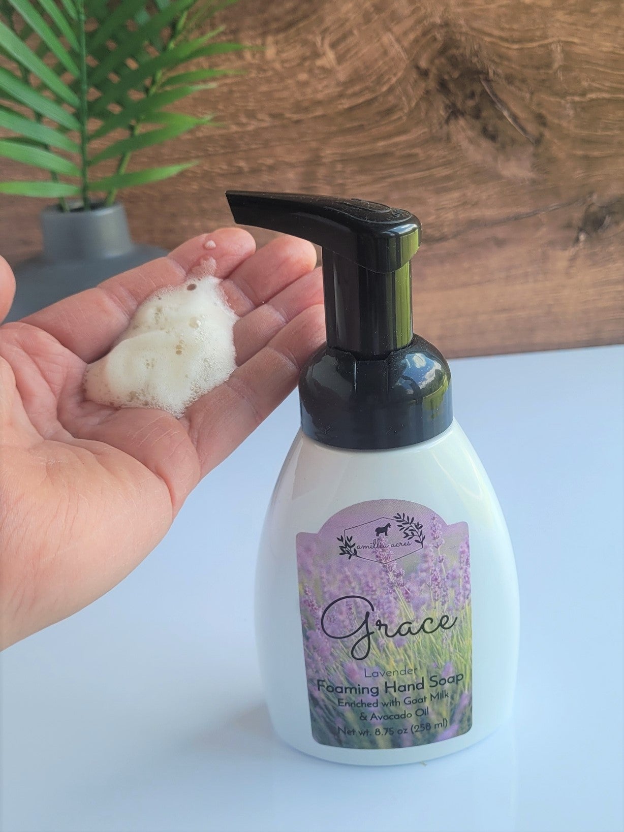 A hand holds "Relax" foaming hand soap from a white Amillia Acres bottle with lavender design against a wooden background and green plant, highlighting the goat milk's moisturizing benefits.