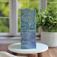 Blueberry Bliss Goat Milk Soap bars by Amillia Acres, featuring blue and silver marbling with glitter accents, are stacked on a white stand. A green potted plant and window appear softly blurred in the background.