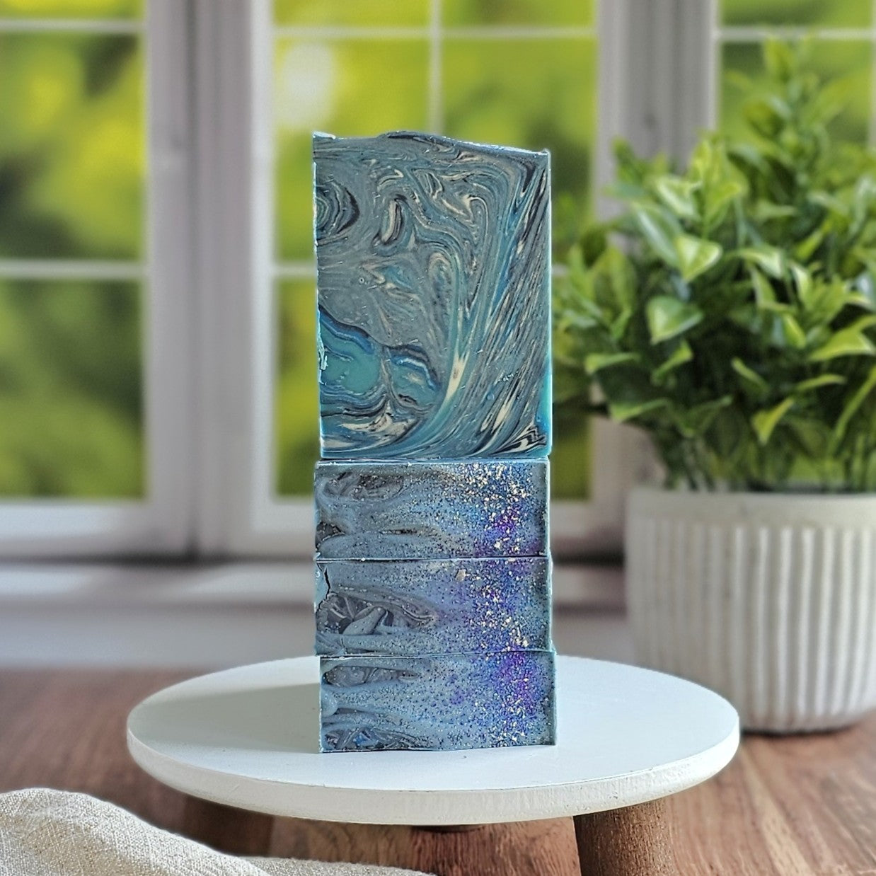 Blueberry Bliss Goat Milk Soap bars by Amillia Acres, featuring blue and silver marbling with glitter accents, are stacked on a white stand. A green potted plant and window appear softly blurred in the background.