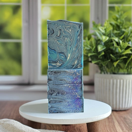 Blueberry Bliss Goat Milk Soap bars by Amillia Acres, featuring blue and silver marbling with glitter accents, are stacked on a white stand. A green potted plant and window appear softly blurred in the background.