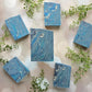Six Blueberry Bliss Goat Milk Soap bars by Amillia Acres are arranged on a light background with bokeh effects, accented by green leaves.