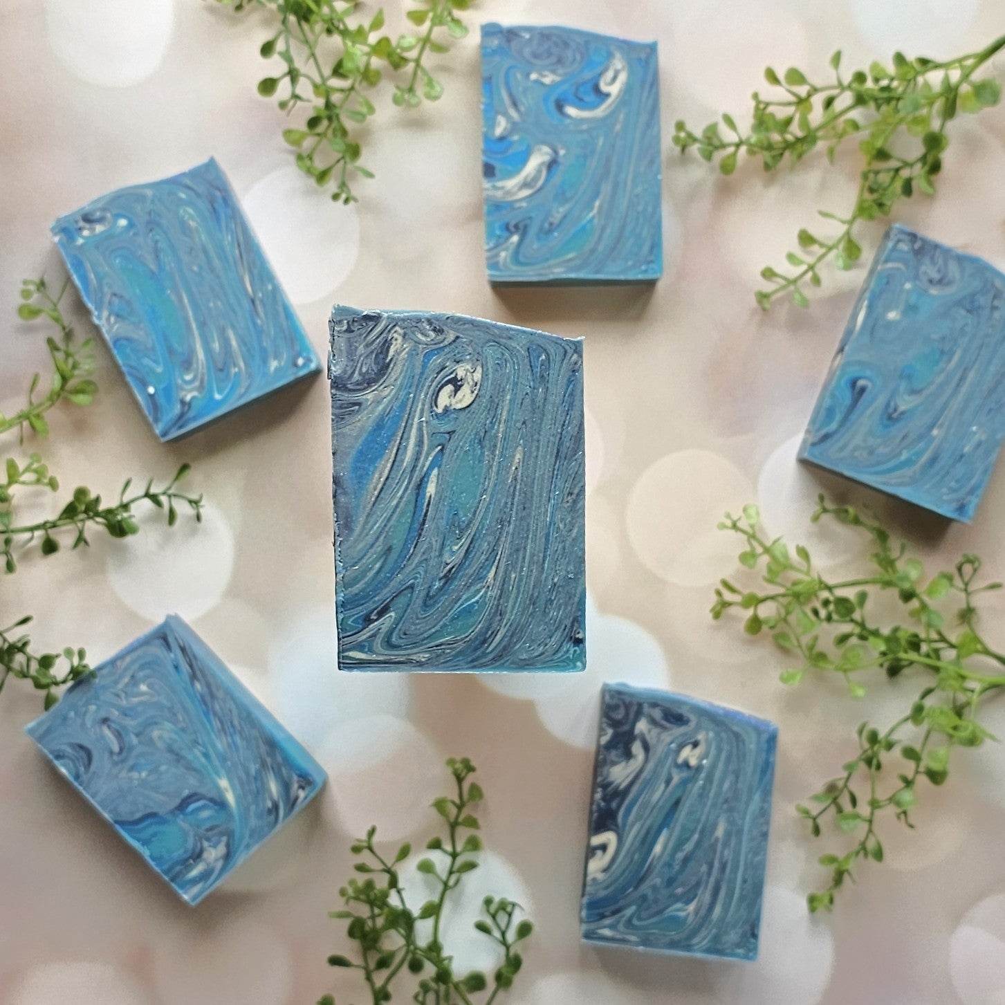 Six Blueberry Bliss Goat Milk Soap bars by Amillia Acres are arranged on a light background with bokeh effects, accented by green leaves.