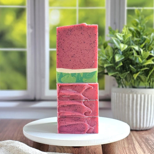 Four pink Melon Baby goat milk soaps by Amillia Acres, with green and beige swirls and exfoliating jojoba beads, are stacked on a round white tray. A potted plant is in the background.