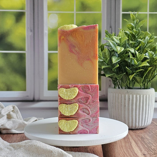 Two Amillia Acres Pink Lemonade Goat Milk Soap bars with pink and yellow swirls and textured yellow-orange segments are stacked on a white stand, in front of a green potted plant and a window showcasing greenery outdoors.