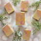 Six bars of Amillia Acres Pink Lemonade Goat Milk Soap are arranged on a light bokeh background, accented with small green leafy sprigs for decoration.