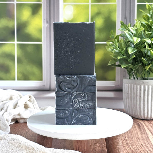 Three rectangular bars of Amillia Acres Balance Goat Milk Soap with marbled dark gray patterns are stacked on a white round stand. A green potted plant and a window with a leafy view appear in the background.