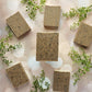 Six rectangular Gardener Goat Milk Soap bars by Amillia Acres, speckled and brown, are arranged in a circle on a light background with small green faux leaves.