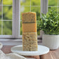 Three stacked Juicy (Essential Oil) Goat Milk Soaps by Amillia Acres sit on a white stand, their varied textures and colors highlighted by natural light. A leafy green plant in a ribbed pot adds freshness beside the handmade bars.