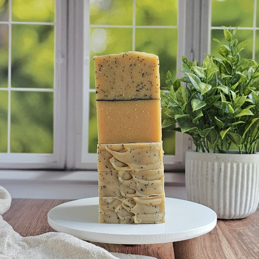 Three stacked Juicy (Essential Oil) Goat Milk Soaps by Amillia Acres sit on a white stand, their varied textures and colors highlighted by natural light. A leafy green plant in a ribbed pot adds freshness beside the handmade bars.