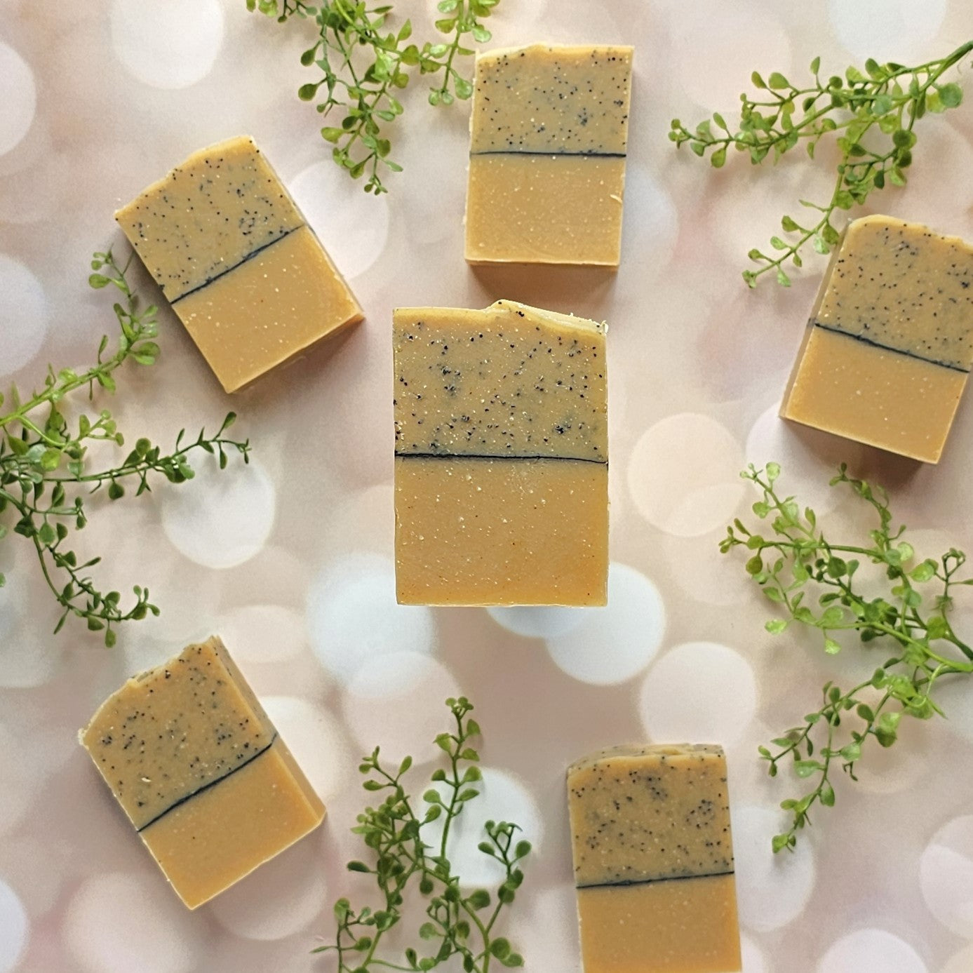Six Juicy (Essential Oil) Goat Milk Soaps by Amillia Acres are arranged on a soft pink bokeh background, each handmade with a speckled top and plain bottom, surrounded by small sprigs of greenery.
