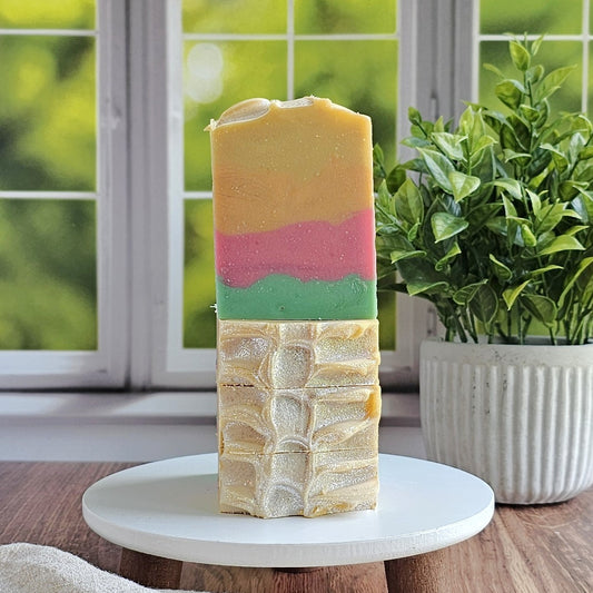 The Amillia Acres Mango Papaya Goat Milk Soap bar features yellow, pink, and green layers. It stands upright on a white round platform with a potted plant and a sunlit window with greenery in the background.