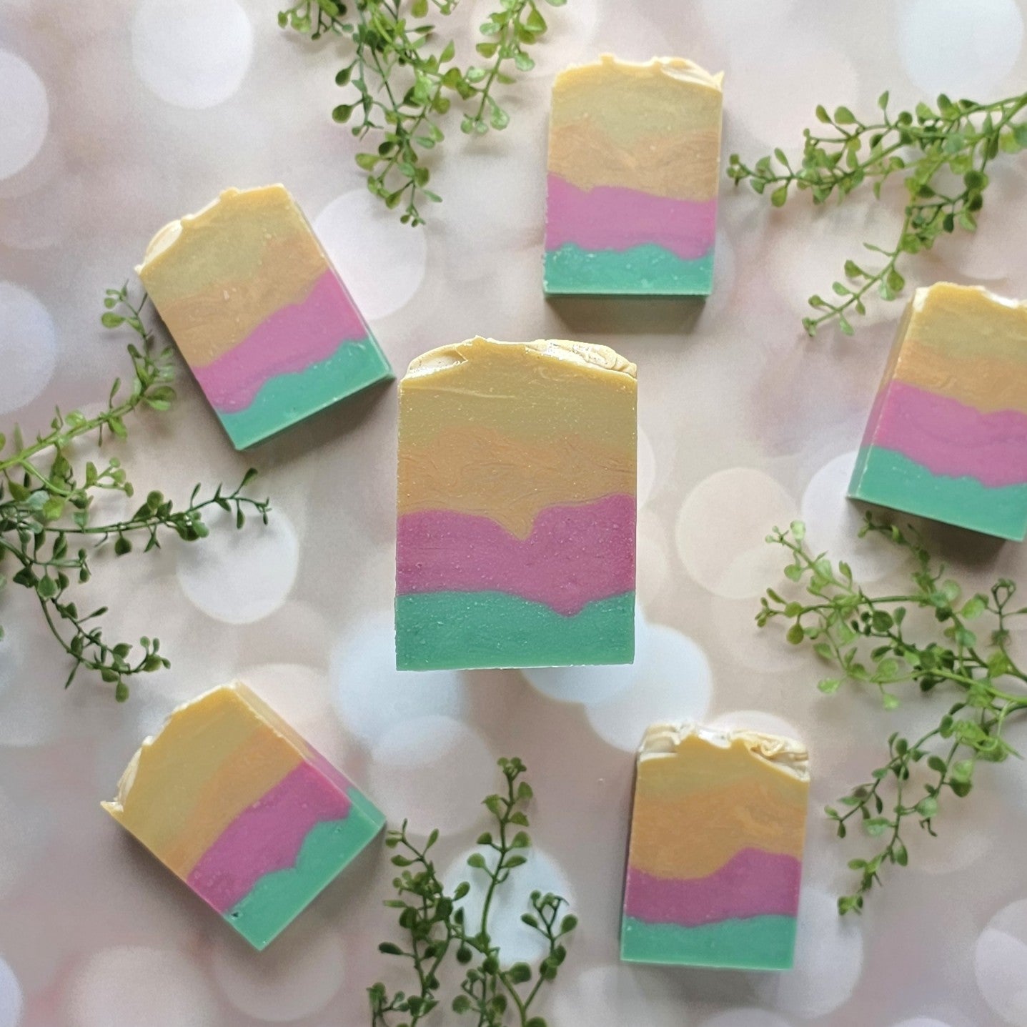 Five pastel Mango Papaya Goat Milk Soap bars by Amillia Acres ~ Goat Milk Soaps and Lotions, each cold pressed with yellow, pink, and teal layers, are arranged in a circle on a soft background with green leafy sprigs.
