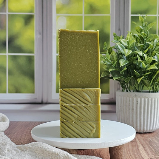 Two green Minty (Essential Oil) Goat Milk Soap bars from Amillia Acres are stacked on a white stand by a window with lush greenery. A plant in a white pot and a beige towel add to the clean, natural feel of the scene.