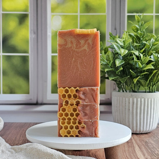 Three rectangular bars of Amillia Acres Rose Clay & Honey Goat Milk Soap are stacked on a white stand. The lower bars show honeycomb patterns and are displayed by a window next to a potted green plant.