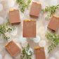 Six Amillia Acres Rose Clay & Honey Goat Milk Soap bars with marbled patterns are arranged on a light, bokeh-patterned surface, accented by green sprigs.