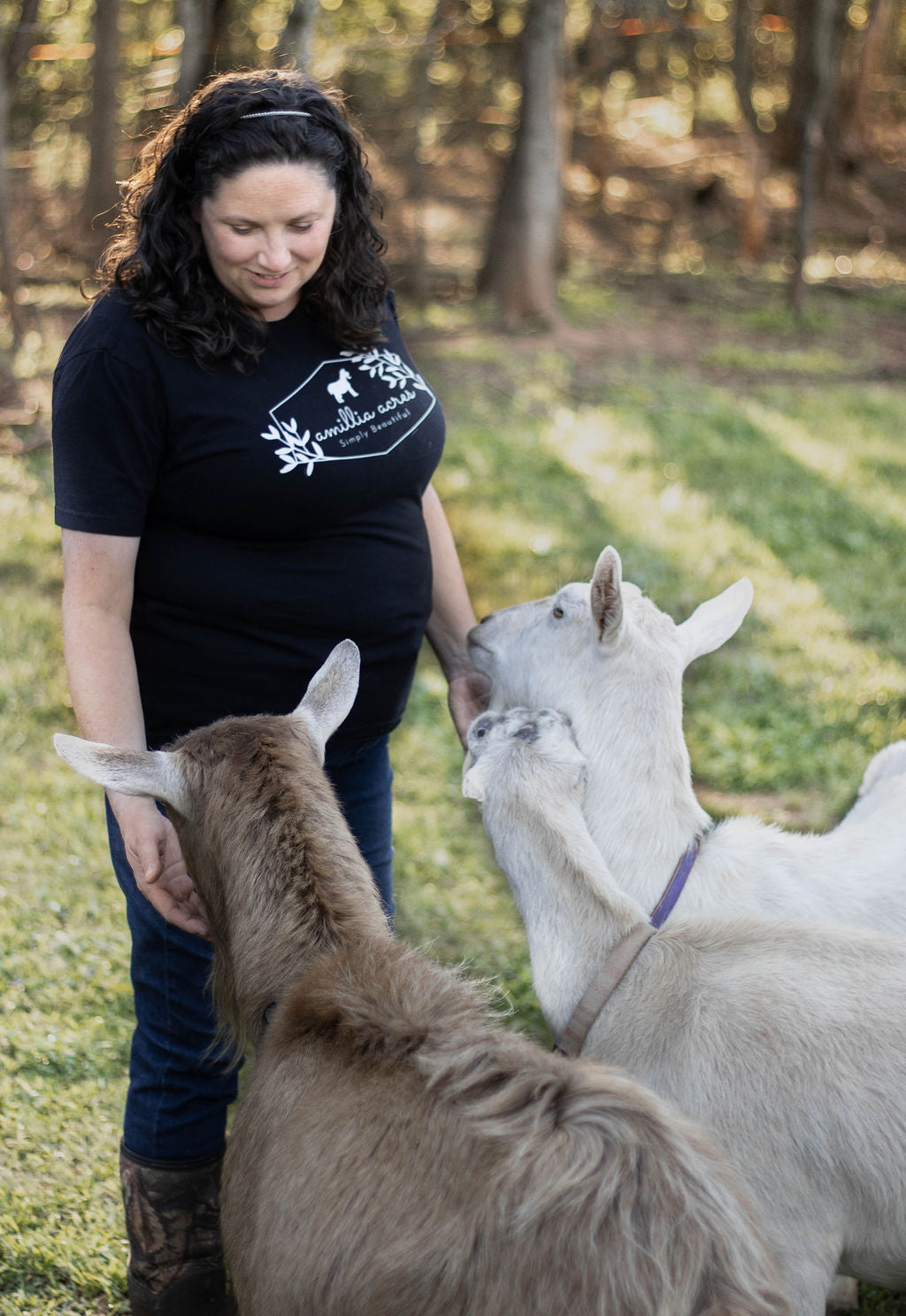 A woman is standing outside, smiling at three goats around her. She is wearing a black t-shirt and jeans. The setting is grassy with trees in the background. The goats are light-colored, and one is nuzzling her hand.