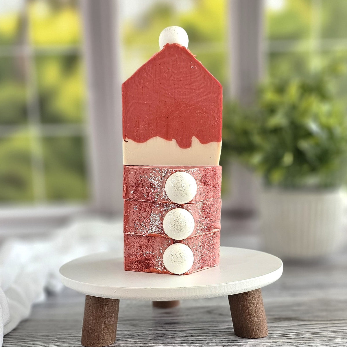 A creatively stacked dessert shaped like a house on a white pedestal resembles the Jolly Goat Milk Soap by Amillia Acres. The top red section mimics a roof, followed by three red layers with white spheres nestled between them, while a blurred green plant adds charm to this scene.