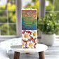 On a small round wooden table stands the Flower Child Goat Milk Soap by Amillia Acres. This handmade bar boasts a colorful rainbow swirl with white daisies on a pink background and is enriched with natural oils, all set against a blurred plant backdrop.