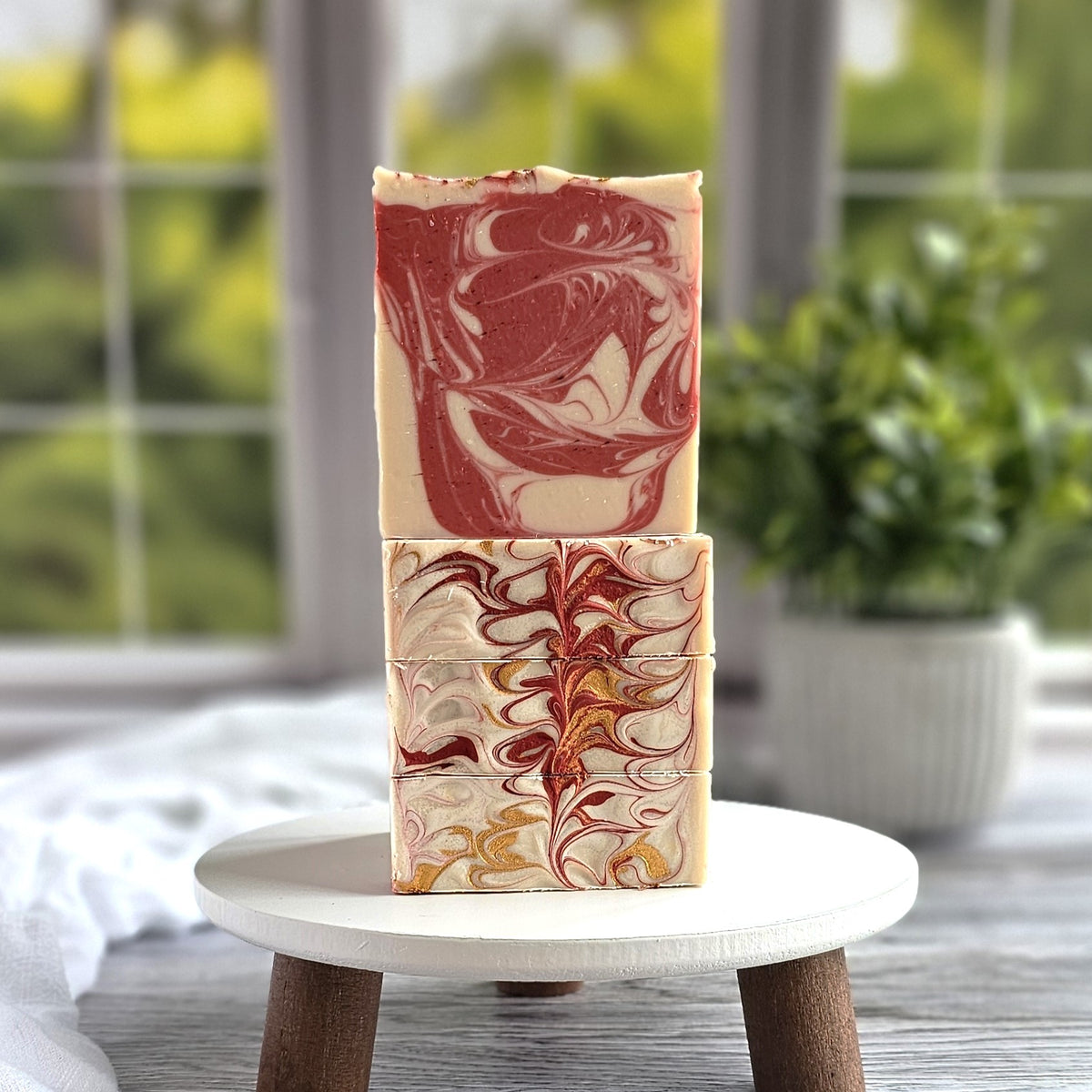 Two Candy Cane Goat Milk Soap bars from Amillia Acres, enriched with natural oils, feature intricate red, yellow, and brown swirls. They rest stacked on a small white round stand with a blurred green plant and window in the background creating a serene atmosphere.