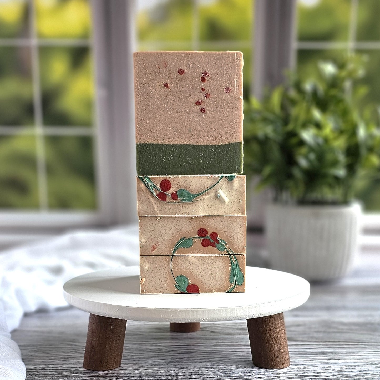 Three Holly Berry Goat Milk Soaps by Amillia Acres are stacked on a round, white wooden stand. Each handmade bar features red and green botanical patterns. A potted plant and a window with greenery outside provide a serene backdrop.