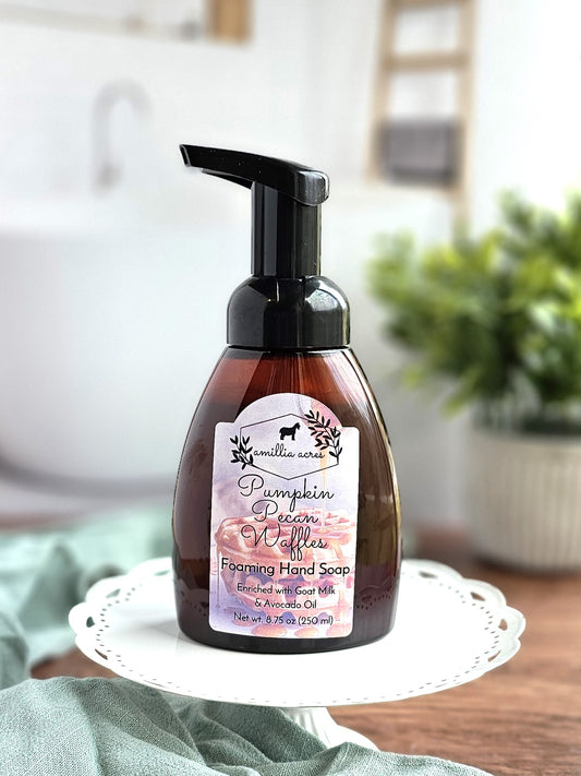 A bottle of Pumpkin Pecan Waffles Foaming Hand Soap by Amillia Acres ~ Goat Milk Soaps and Lotions rests on a white decorative stand. The label displays autumn-themed graphics, and in the background, a hint of a bathroom setting with a potted plant adds to the cozy scene.