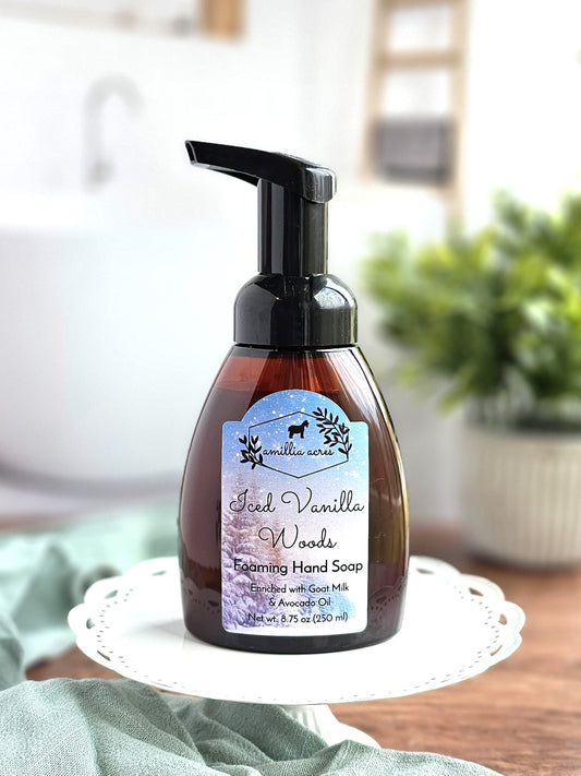 A bottle of Iced Vanilla Woods Foaming Hand Soap from Amillia Acres ~ Goat Milk Soaps and Lotions rests on a white decorative tray. The label showcases a wintry forest design. In the background, there's a blurry bathroom setting with a plant and white basin.