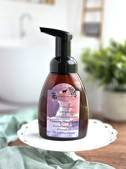 A pump bottle of Amillia Acres' Vanilla Pumpkin Marshmallow Foaming Hand Soap, enriched with moisturizing goat milk, is elegantly placed on a white decorative stand. The label features autumnal colors and designs. In the softly blurred background, hints of greenery and household items are visible.