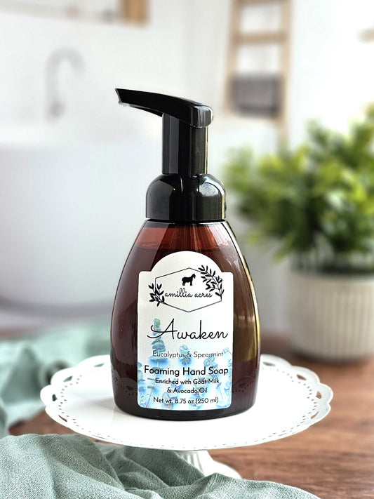 A bottle of Amillia Acres' "Awaken" Foaming Hand Soap (Eucalyptus & Spearmint) is elegantly displayed on a white stand. Enriched with goat milk and avocado oil, the soap features refreshing notes of eucalyptus and spearmint. In the blurred background, plant and kitchen items add a natural charm to the scene.