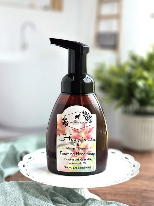 A bottle of Amillia Acres' "Happiness" Foaming Hand Soap, featuring a mango tangerine scent, is elegantly displayed on a white decorative stand. The label emphasizes its enrichment with goat milk and avocado oil. In the background, a blurred bathroom setting enhanced by touches of greenery complements the scene.