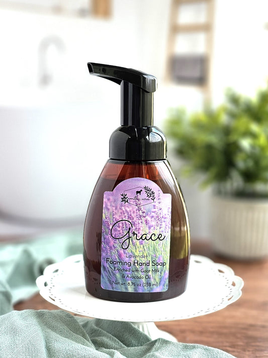 A bottle of Grace Foaming Hand Soap from Amillia Acres ~ Goat Milk Soaps and Lotions, featuring a lavender-themed label, sits on a white decorative stand. In the background, a blurred sink and a green potted plant create a clean and fresh bathroom setting.