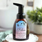 A pump bottle of Amillia Acres' "Relax" Foaming Hand Soap, featuring jasmine and driftwood on the label, enriched with goat milk and avocado oil. It sits elegantly on a decorative white plate with a softly blurred plant in the background.