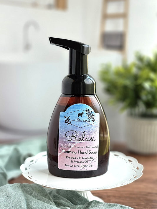 The "Relax Foaming Hand Soap" by Amillia Acres ~ Goat Milk Soaps and Lotions is a moisturizing hand soap in a pump bottle, infused with calming jasmine and driftwood scents, along with nourishing goat milk and avocado oil. It is beautifully displayed on a decorative tray next to a plant against a softly blurred white backdrop.