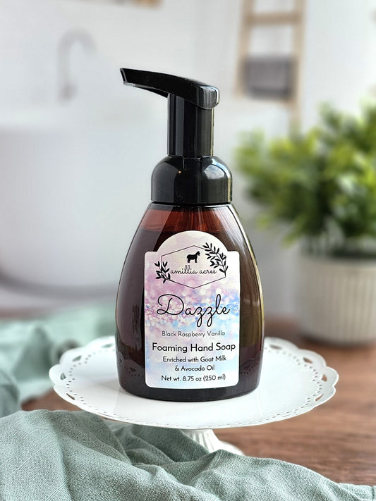 A bottle of Amillia Acres ~ Goat Milk Soaps and Lotions Dazzle Foaming Hand Soap (Black Raspberry Vanilla) sits elegantly on a white cake stand. The background features a blurred indoor setting decorated with a plant and towel.