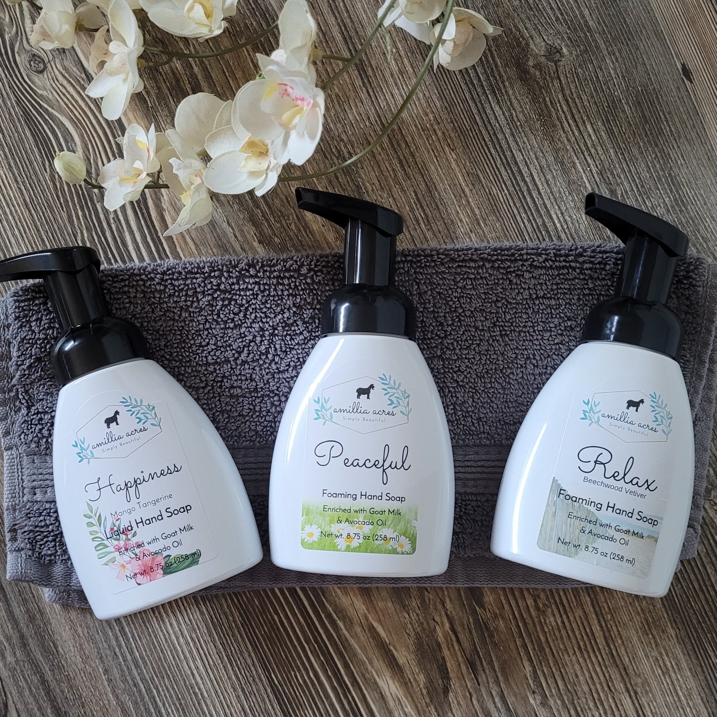 Three bottles of Amillia Acres's Comfort Foaming Hand Soap, scented with Apricot & Honey and labeled "Happiness," "Peaceful," and "Relax," sit on a gray towel. Each bottle features floral and natural designs, capturing the essence of moisturizing hand soap. A branch of white orchids accents the wooden surface.