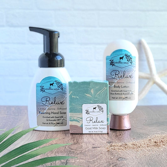 The "Relax Signature Bundle" from Amillia Acres ~ Goat Milk Soaps and Lotions, is showcased on a wooden surface. This collection includes goat milk foaming hand soap and body lotion, all adorned with blue and white labels. Delight in calming bestseller scents such as coconut, jasmine, and driftwood. A plant is partially visible in the background.