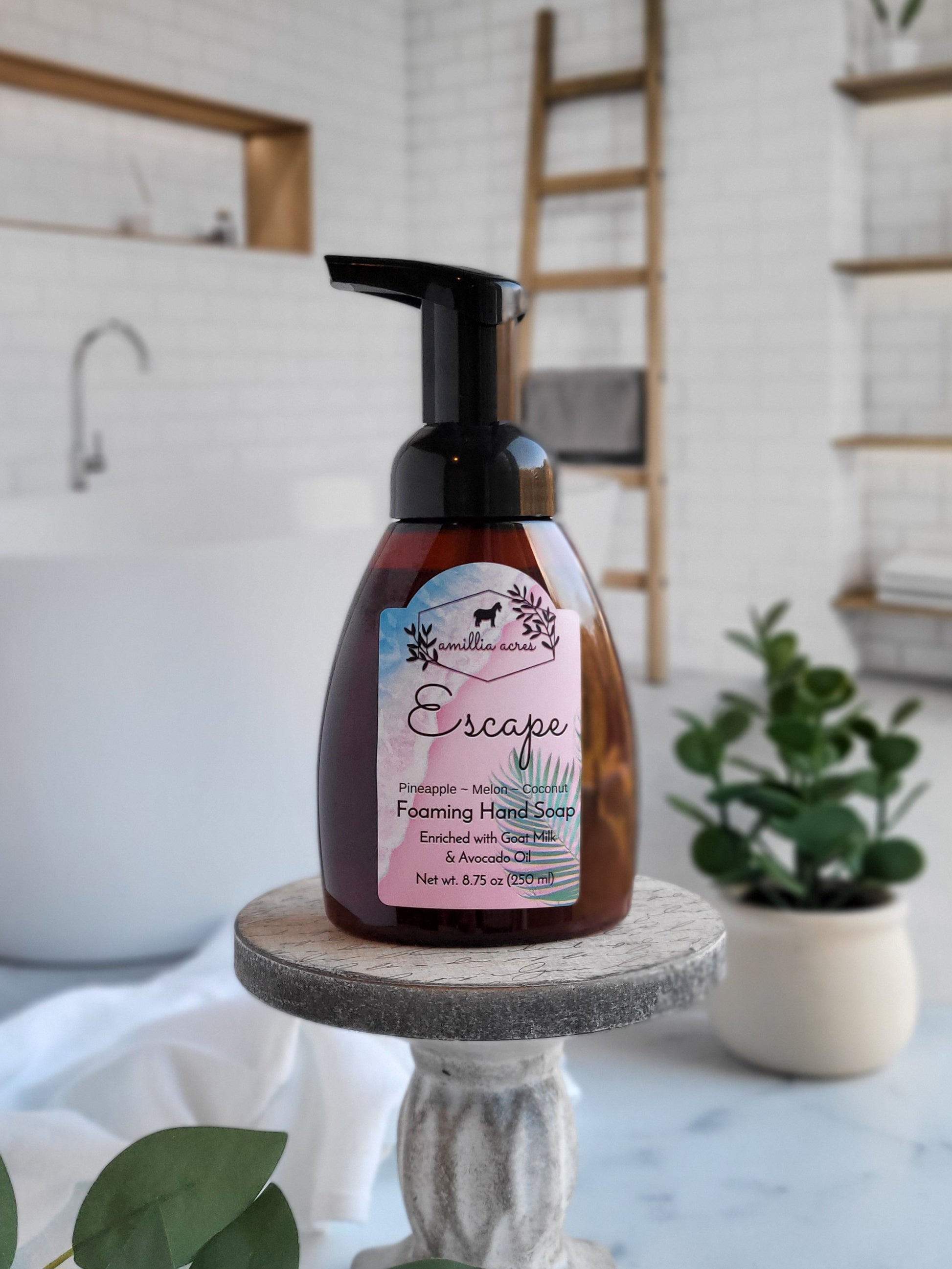 A bottle of Amillia Acres' Escape Foaming Hand Soap, featuring a pineapple and melon scent, sits on a pedestal. The scene includes a modern bath with a white tub, plants, and wooden ladder—an ideal setting for your moisturizing hand care routine.