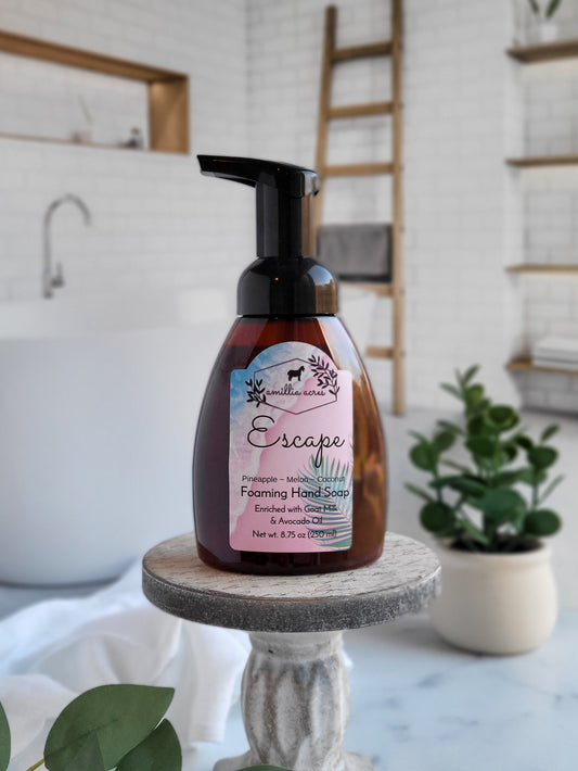 An "Escape Foaming Hand Soap" bottle from Amillia Acres ~ Goat Milk Soaps and Lotions, featuring a tropical design, sits on a pedestal in a bright bathroom. In the background, you can see a bathtub, plants, and a wooden ladder that together create a spa-like atmosphere.