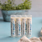 Four tubes of Simply Natural Lip Balm by Amillia Acres ~ Goat Milk Soaps and Lotions, adorned with blue floral designs, stand upright on a light blue surface. Enriched with a nourishing recipe full of antioxidants, they promise to deliver soft lips. A blurred green plant in the background adds a touch of nature to the scene.