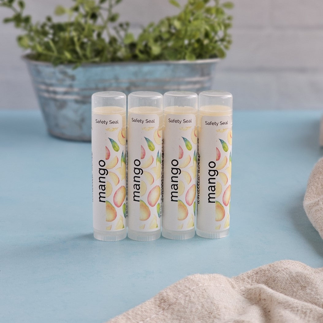 Four tubes of Amillia Acres' Mango Lip Balm, enriched with nourishing butters, are arranged on a light blue surface, featuring mango illustrations. In the blurred background sits a metal pot with green plants, and a beige cloth is partially visible on the right.