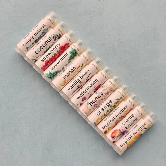 flavors of lip balms in a row - there are simply natural, coconut, strawberry, spearmint, mango, vanilla bean, watermelon, honey, orange, tropical medley, creme brulee, and tropical paradise pictured here
