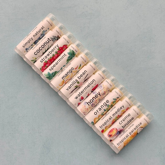 lip balms lined up at a diagonal on a blue background - pictured here are simply natural, coconut, strawberry, spearmint, mango, vanilla bean, watermelon, honey, orange, tropical medley, creme brulee, tropical paradise lip balms.