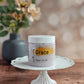 A jar of Amillia Acres ~ Goat Milk Soaps and Lotions Grace Sugar Scrub rests on a white decorative cake stand atop wood, highlighting this luxurious exfoliating body scrub with a blurred floral vase in the background.