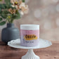 A jar of Dazzle Sugar Scrub by Amillia Acres ~ Goat Milk Soaps and Lotions in Black Raspberry Vanilla rests on a white cake stand on a wooden surface, with blurred flowers visible in the background.