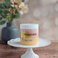 A jar of Happiness Sugar Scrub by Amillia Acres ~ Goat Milk Soaps and Lotions sits on a white cake stand, offering glowing skin with its exfoliating blend; a blurred vase of flowers rests in the background on a wooden surface.
