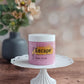A pink jar of Amillia Acres ~ Goat Milk Soaps and Lotions "Escape | Sugar Scrub" exfoliating body scrub for glowing skin sits on a white cake stand, with a blurred gray vase of flowers in the background on a wooden surface.